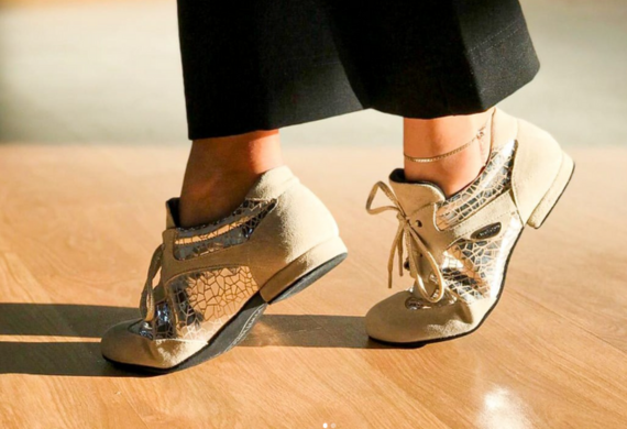 Person, die Tanzschuhe mit beigem und metallischem Netzdesign trägt und im Sonnenlicht auf einem Holzboden steht.